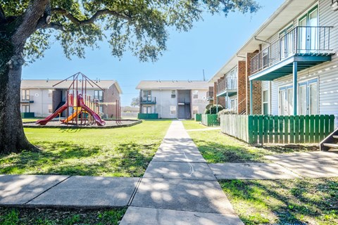 A playground with a slide is in the middle of a grassy area between two apartment buildings.