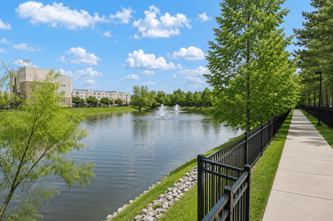 A serene lake with a fountain in the middle, surrounded by a black fence and green trees.
