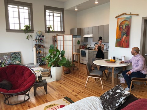 A man is sitting at a table in a room with a woman standing in the kitchen.
