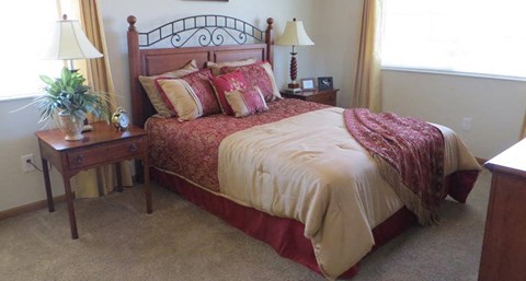 A bed with a red and white comforter and pillows sits in a bedroom with a nightstand and a plant.