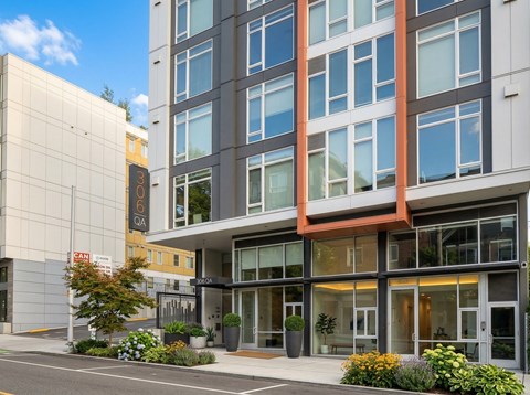 Street-level entrance of 306 QA apartment building with modern glass doors, potted plants, and sidewalk flower beds.