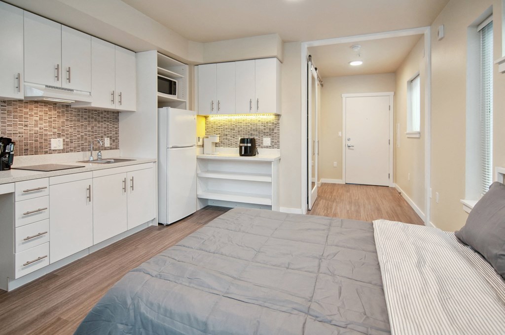 A bedroom with a bed, a kitchen area, and a hallway.