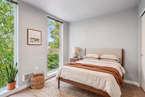 Bedroom with a queen bed, wood nightstand and two full-height windows looking into leafy tree cover.
