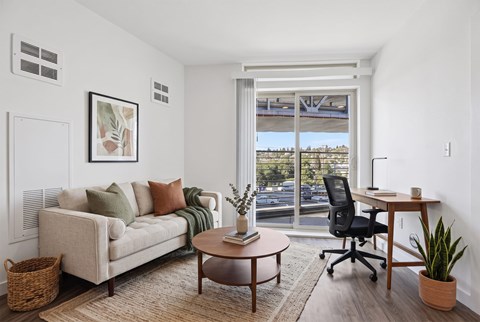 Living area with a sofa, round coffee table, wood desk and task chair beside a sliding glass door to the balcony.