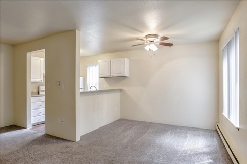 an empty living room with a ceiling fan and a kitchen in the background