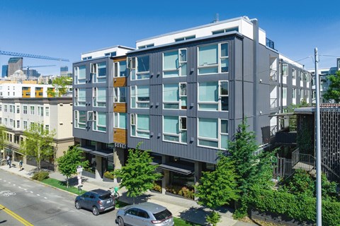 A modern building with a grey facade and large windows is surrounded by trees and parked cars.
