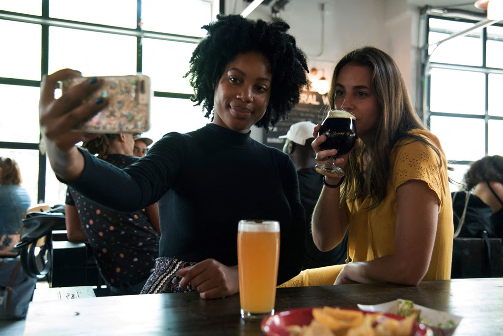 two women sitting at a table drinking beer and looking at a cell phone