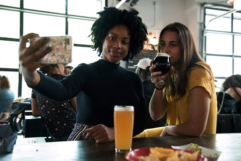 two women sitting at a table drinking beer and looking at a cell phone
