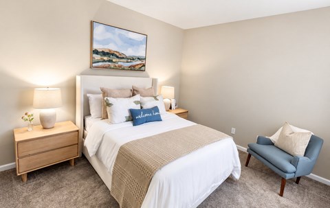 A bedroom with a large bed, a nightstand with a lamp, a chair, and a painting on the wall.