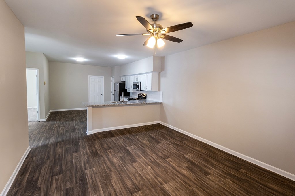 an empty living room with a ceiling fan and a kitchen in the background