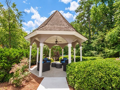 A gazebo with a brown roof is surrounded by green bushes and trees.