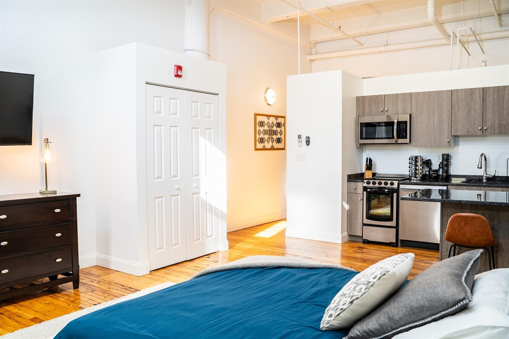 A bedroom with a bed, dresser, and a kitchen area in the background.