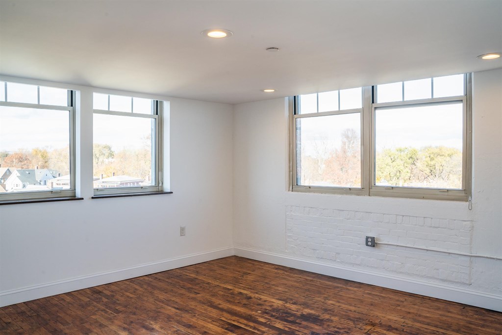 A room with white walls and wooden floors with two windows.