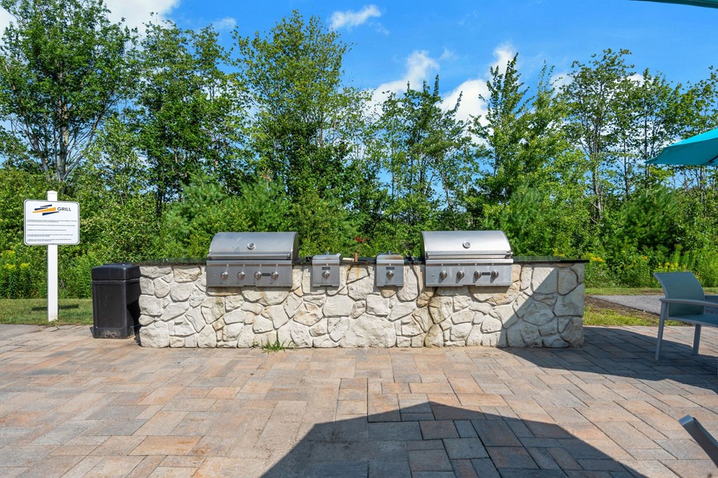 a stone wall with three barbecue pits and trees in the background at The Beacon at Gateway Apartments, Scarborough, ME, 04074