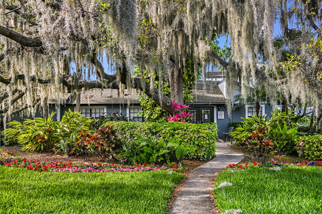View of Brookside Manor Clubhouse and walkway with vivid green grass and red and pink foliage, underneath a mature tree canopy