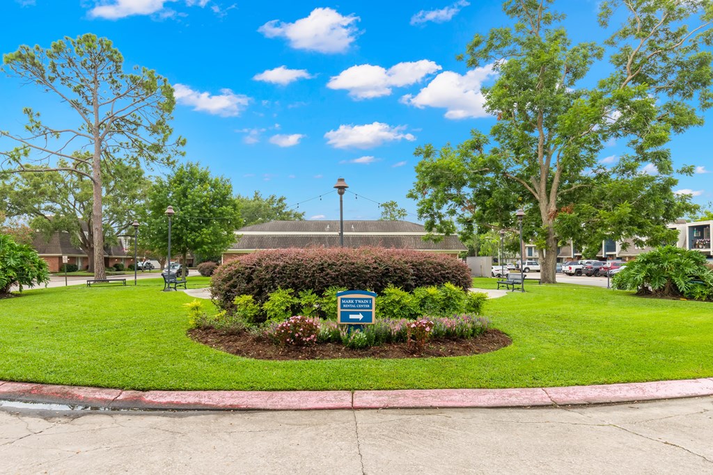 A sign in a grassy area with a tree and a building in the background.