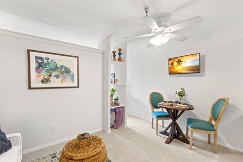 A room with a ceiling fan, a painting on the wall, a small table with chairs and a basket on the floor.