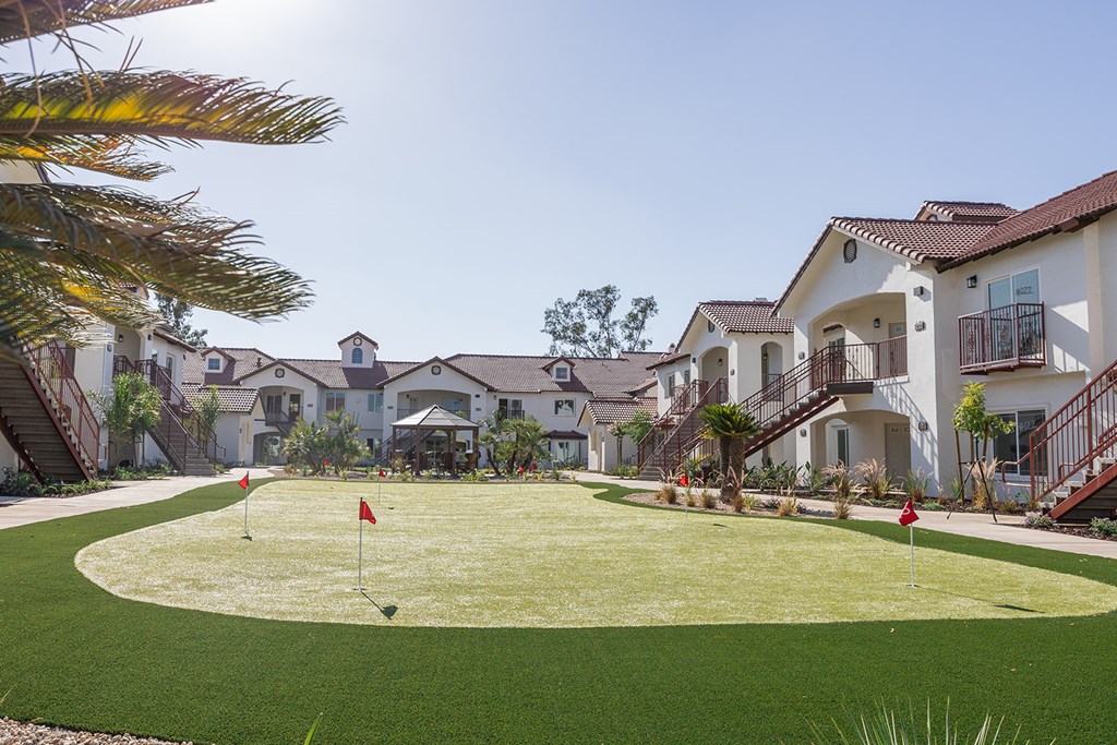 A golf hole with a red flag on a sunny day in front of a row of houses.