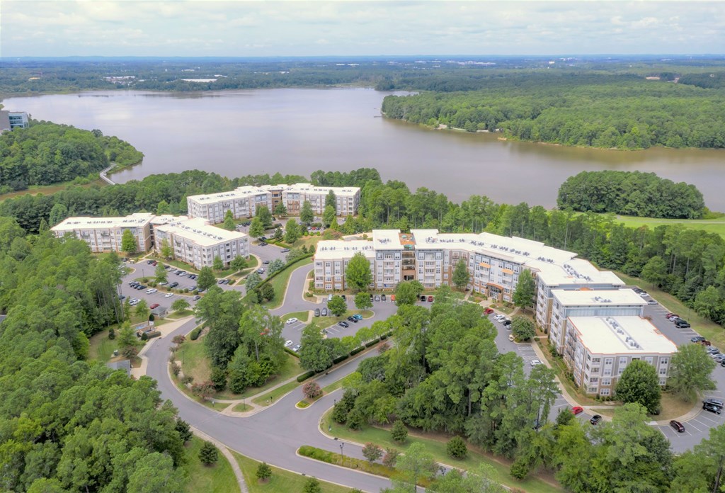 A large white building complex is surrounded by trees and a body of water.