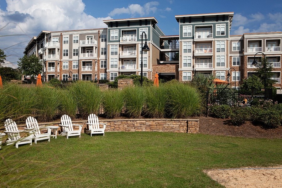 A row of white chairs are set up on a lawn in front of apartment buildings.