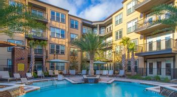 A large swimming pool is surrounded by lounge chairs and palm trees in front of apartment buildings.