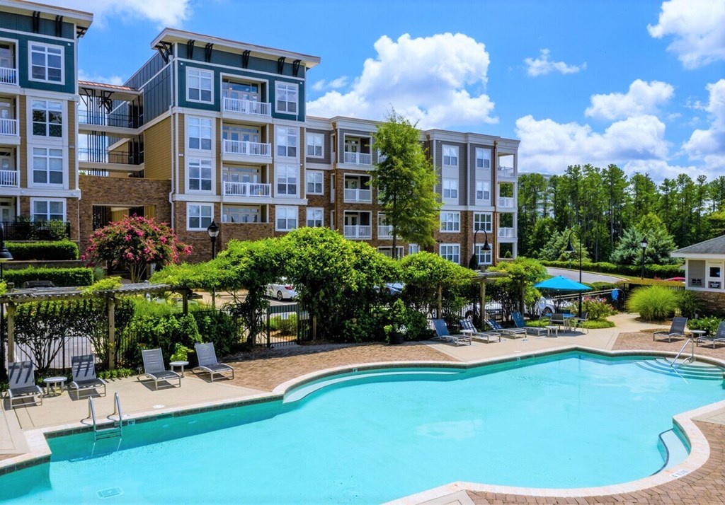 A swimming pool surrounded by lounge chairs and trees in front of apartment buildings.