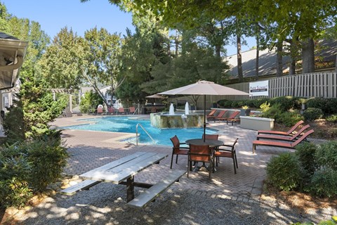 A pool surrounded by trees and a patio with chairs and an umbrella.