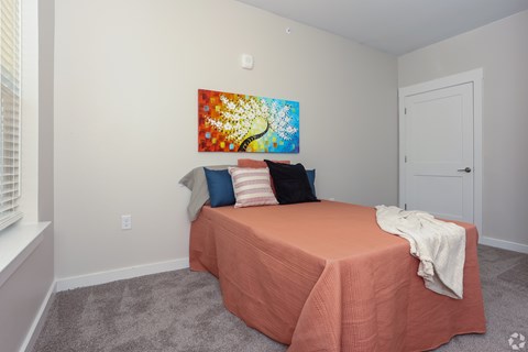 A bedroom with a bed covered in a peach sheet and a colorful painting on the wall.