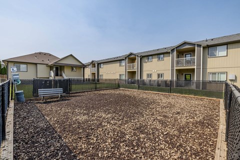 A playground area with a brown surface surrounded by a black fence in front of apartment buildings.