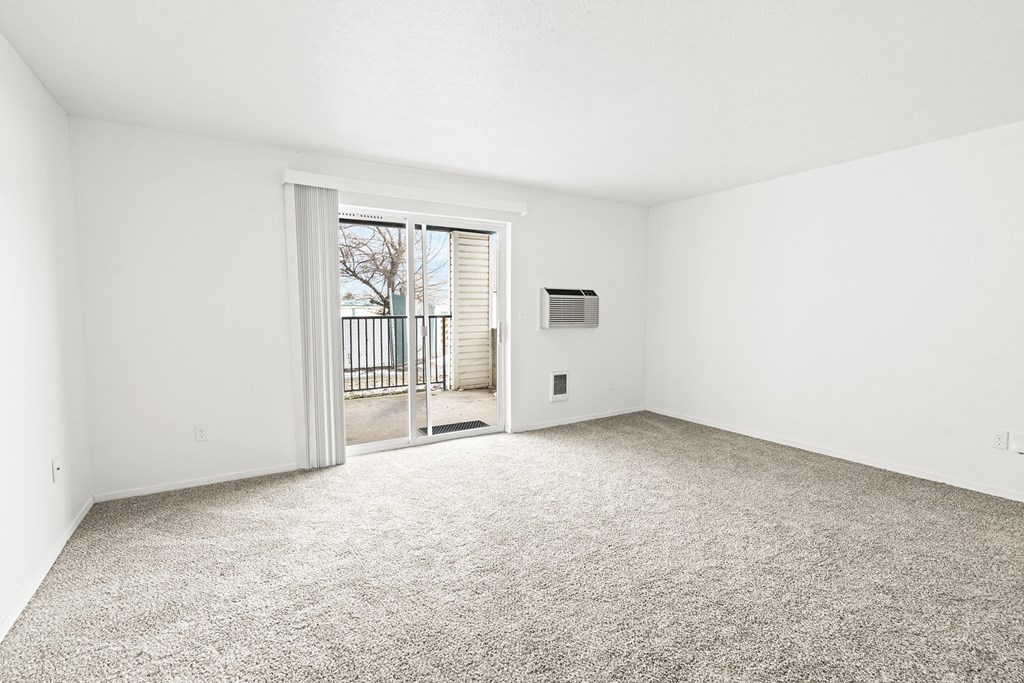 Bright white living room area with taupe carpet, and sliding glass door with vertical blinds at far end of the room..at North Pointe, Post Falls, 83854