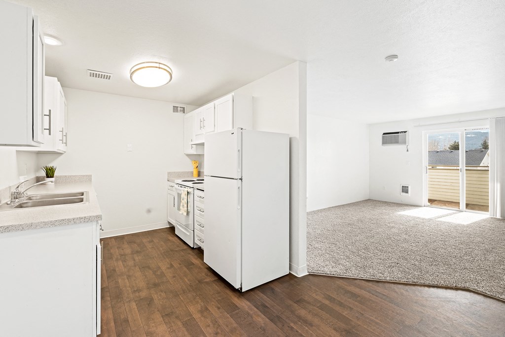 White kitchen with new cabinets, dark faux wood flooring, and view of living room area with taupe carpet and sliding glass door in the distance. at North Pointe, Post Falls, ID 83854