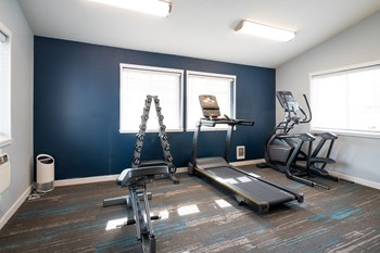 Fitness center with white walls and one dark blue accent wall. An elliptical machine, treadmill and stand with free weights and weight bench are visible.. - Photo Gallery 10