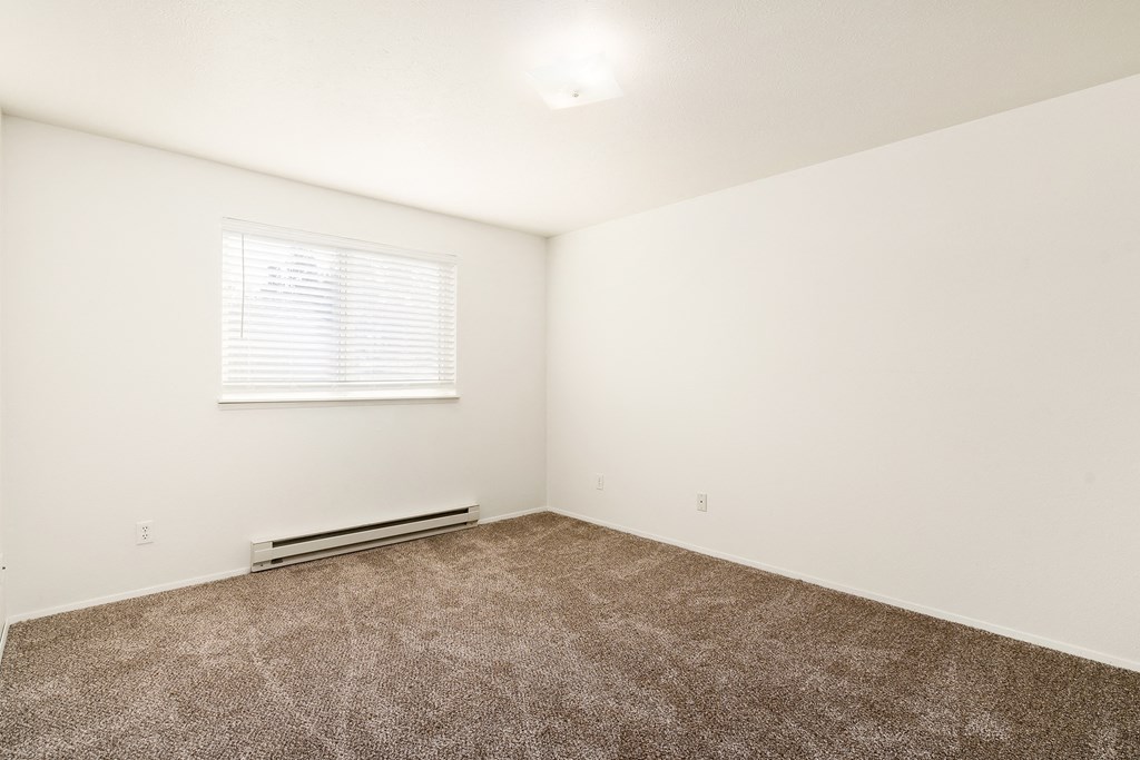 A spacious bedroom with white walls, a large window with blinds, and taupe carpets.at Clearwater, Post Falls Idaho