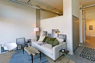 Apartment living room with staged couch, coffee table and rug. Concrete flooring and tall ceilings.