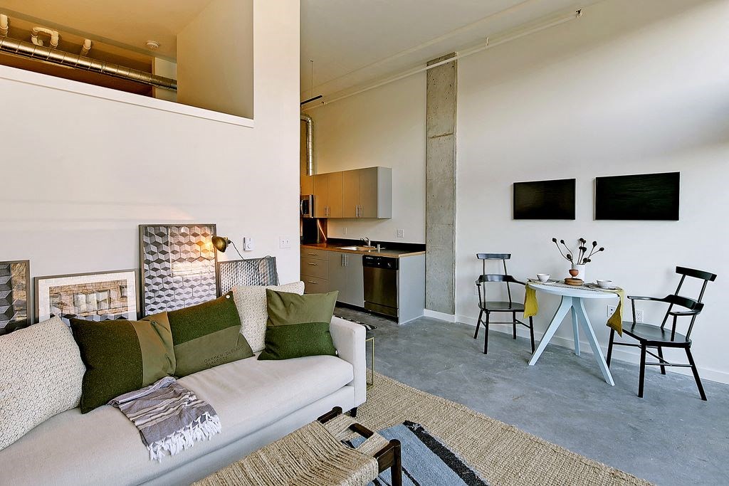 Living room with partial wall behind couch, leading to loft area. Staged with a couch, coffee table, rug and chair. Small dining area at right, with kitchen behind.at 19th & Mercer, Seattle Washington