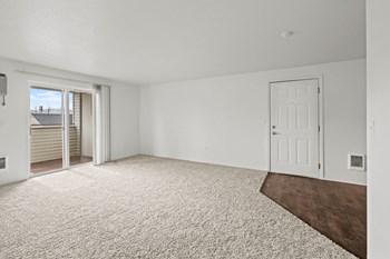 Bright white living room area with taupe carpet, and sliding glass door with vertical blinds at far end of the room. View of the white front door and dark brown faux wood flooring at entry. - Photo Gallery 18