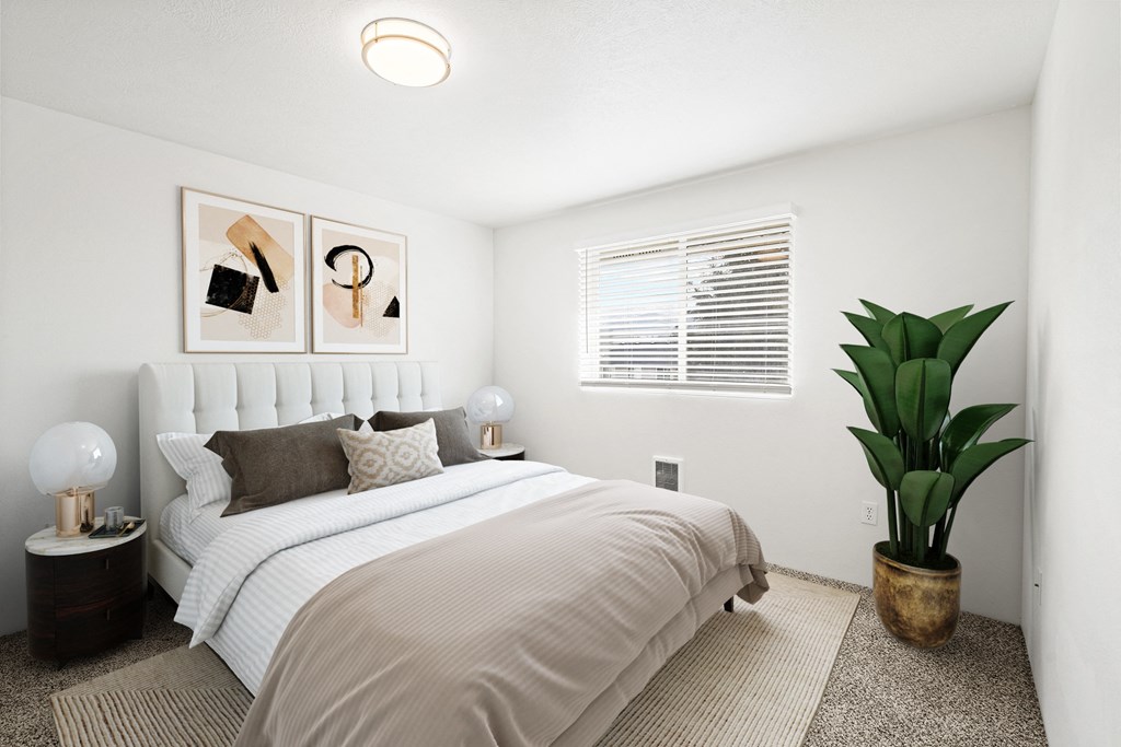 a large bedroom with white walls, taupe carpet, and a window on the back wall. Staged with queen-size bed, 2 side tables, art and a plant.at North Pointe, Post Falls