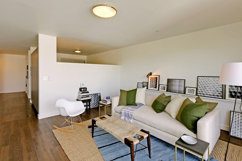 Living room wit partial wall into bedroom area. Hallway visible with build-in storage. Staged with a couch, coffee table, rug and chair.at 19th & Mercer, Seattle Washington