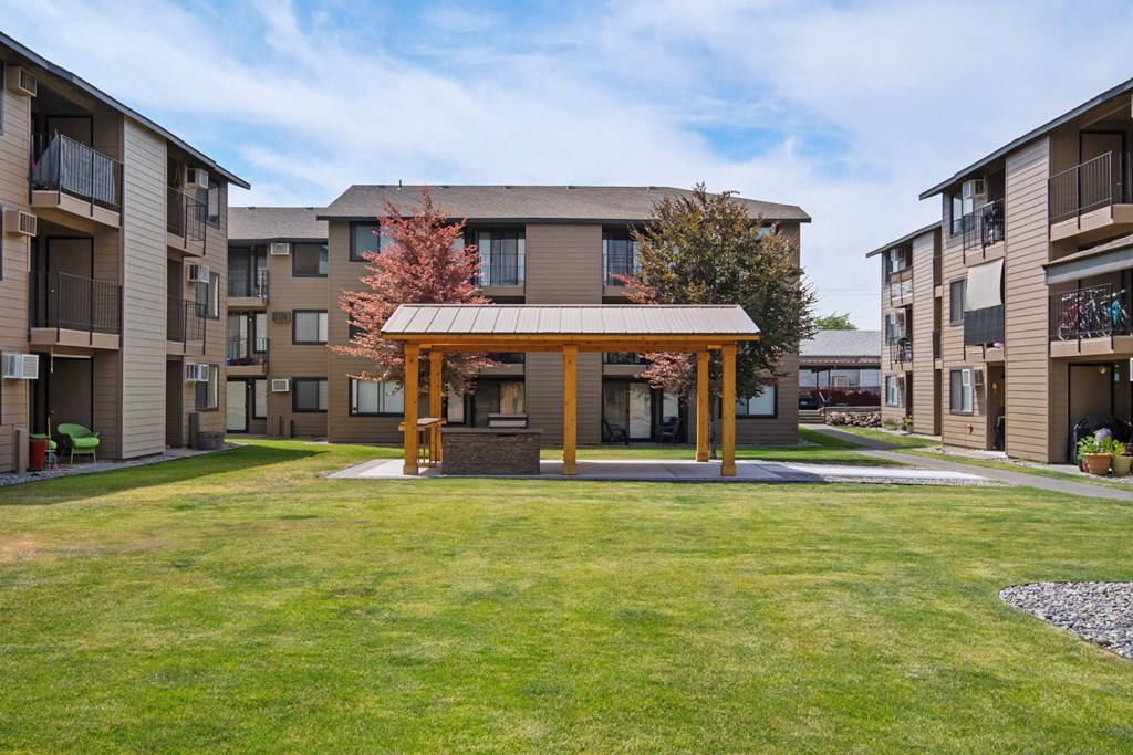 The Brix community courtyard surrounded by grass, trees, and apartments with a covered structure for residents in the center. at Brix, Washington, 99362
