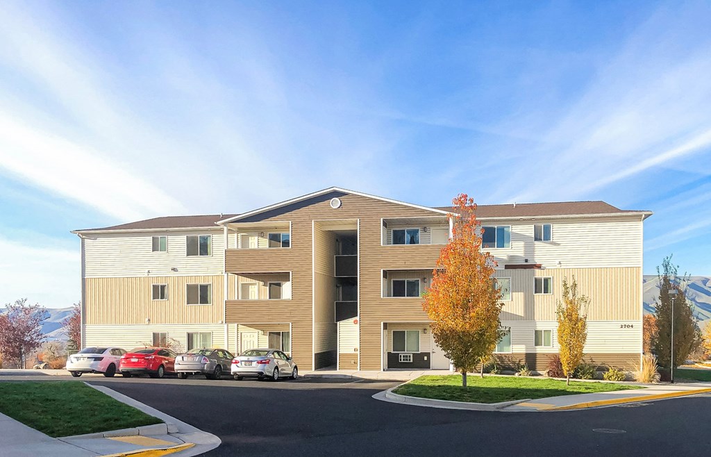 Exterior of Canyon View Apartments with mountains and blue skies behind, parked cars to the left, and landscaping on the right.at Canyon View, Lewiston, ID.