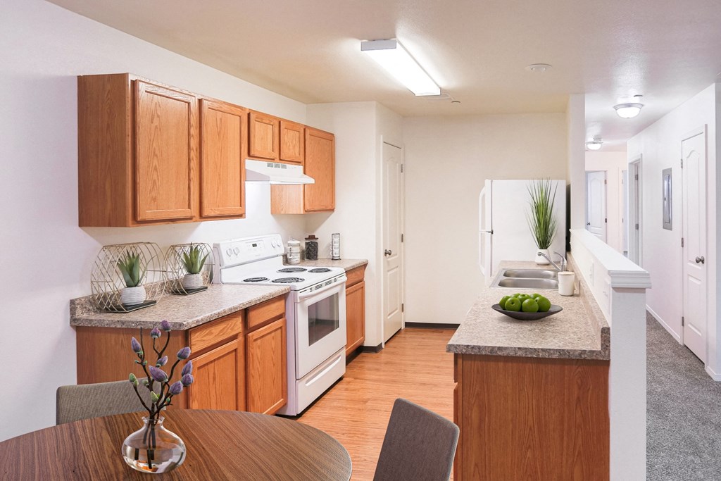 Bright kitchen with honey-colored cabinets and white appliances and faux wood flooring. You can also see down the hallway with white walls and taupe carpet.at Canyon View, Lewiston