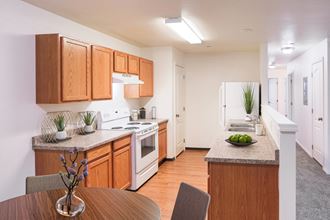 Bright kitchen with honey-colored cabinets and white appliances and faux wood flooring. You can also see down the hallway with white walls and taupe carpet.