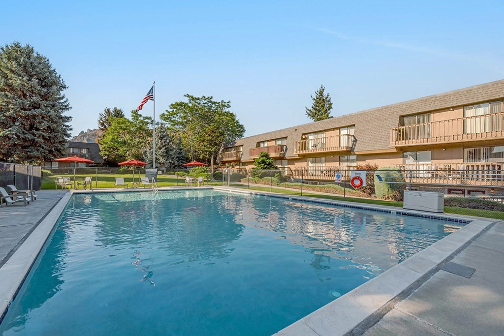 Large swimming pool, lounge chairs, tables, umbrellas, enclosed with fence surrounded by grass and apartments with balconies.at Castlerock, Washington