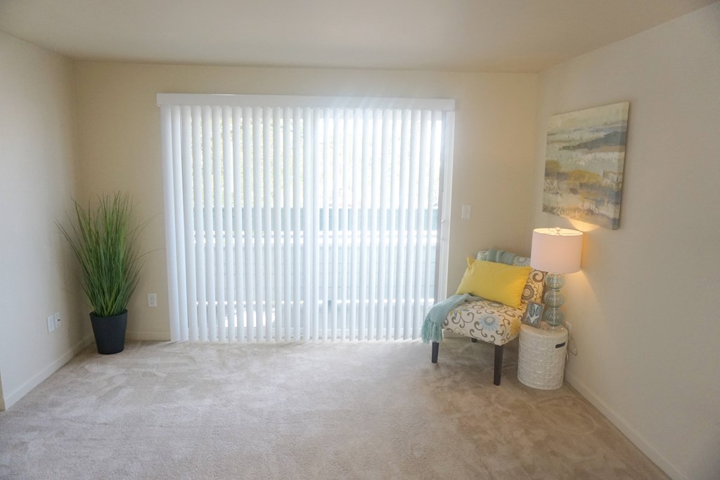 a living room with a large sliding glass door, beige carpet and a chair at Heritage Grove Apartments in Renton, WA