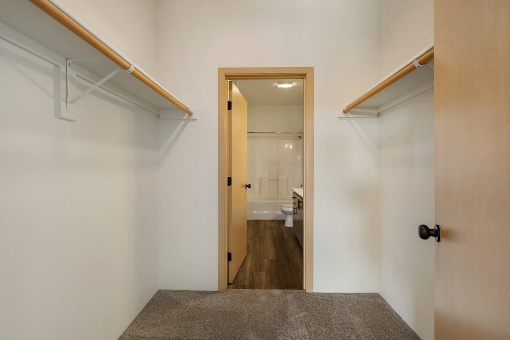 A walk-in closet with custom shelving, taupe carpets, white walls and access to the bathroom.at Shoreline Village, Richland, 99352