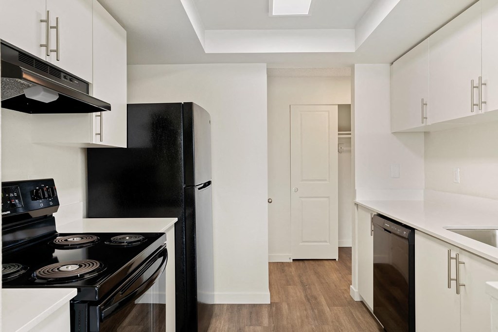 A galley-style kitchen with white cabinets, black appliances, and access to the hallway and hallway closet behind. at Pointe East, Fife, WA, 98424