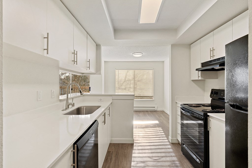 Light-filled galley kitchen with white countertops and cabinets looks into the dining room with sunny windows and white walls. at Pointe East, Fife, WA