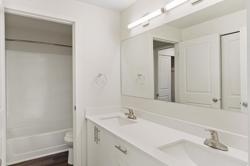 A full bathroom with double vanity sinks, white countertops and cabinets, a large mirror, and a door to the toilet and shower. at Pointe East, Fife, 98424