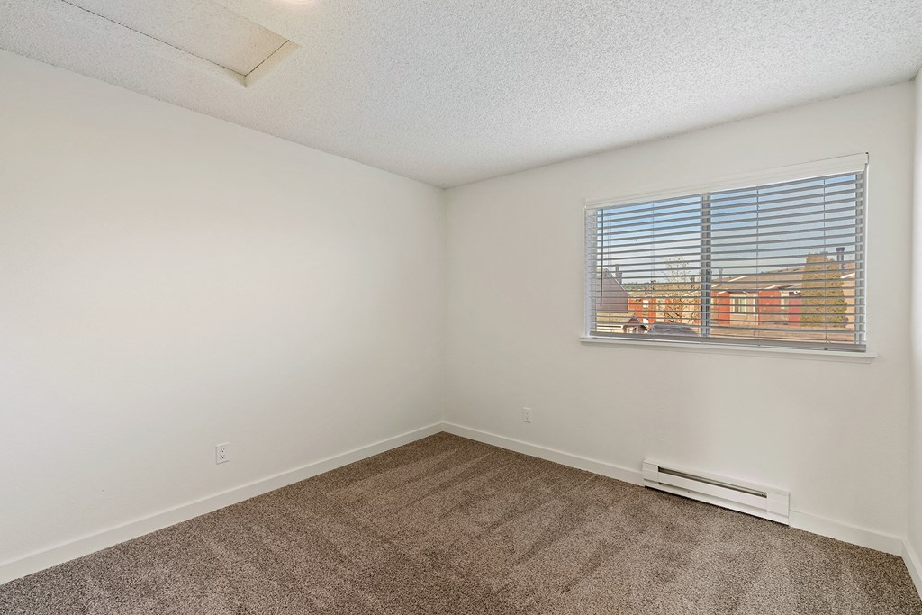 A bedroom with white walls, taupe carpets, a double window with white blinds, and a view on a sunny day. at Pointe East, Fife, Washington