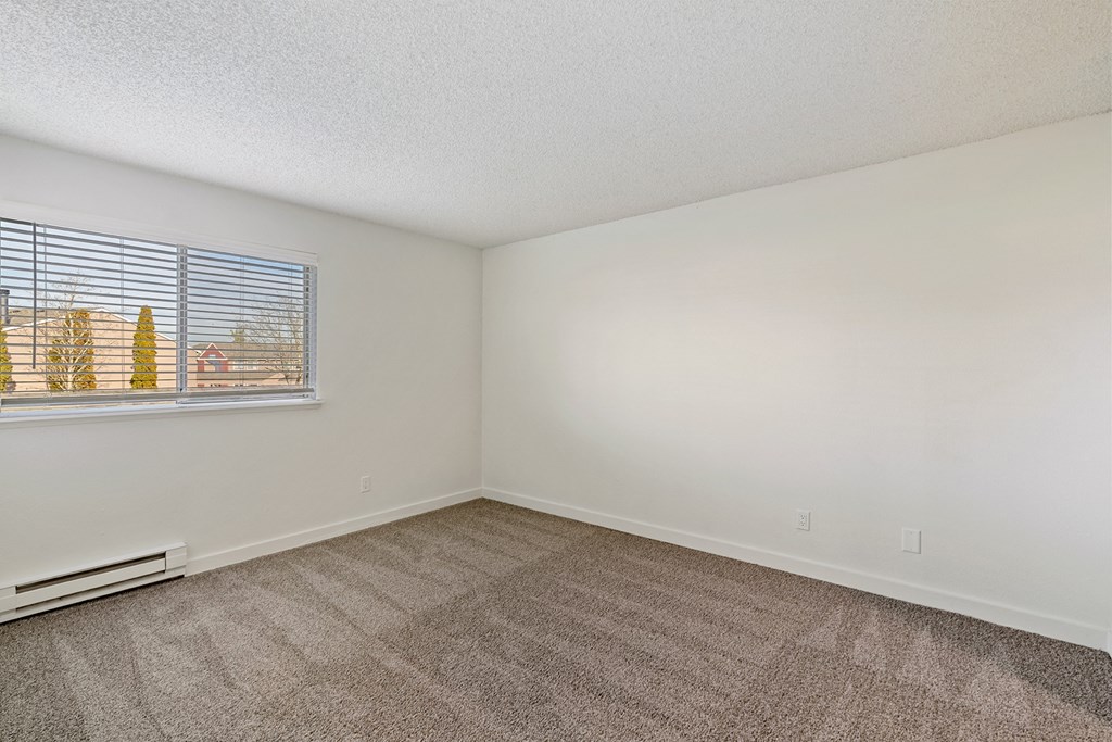 A spacious bedroom with white walls, sunny windows, a heater below the window, and taupe carpets. at Pointe East, Washington, 98424